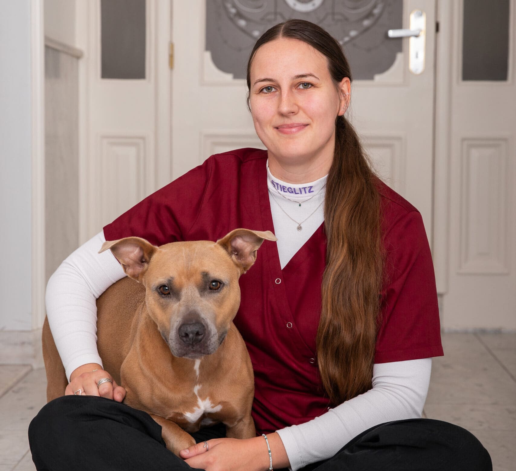 Medewerker in bordeauxrode dienstkleding zit op de vloer naast een lichtbruine hond.