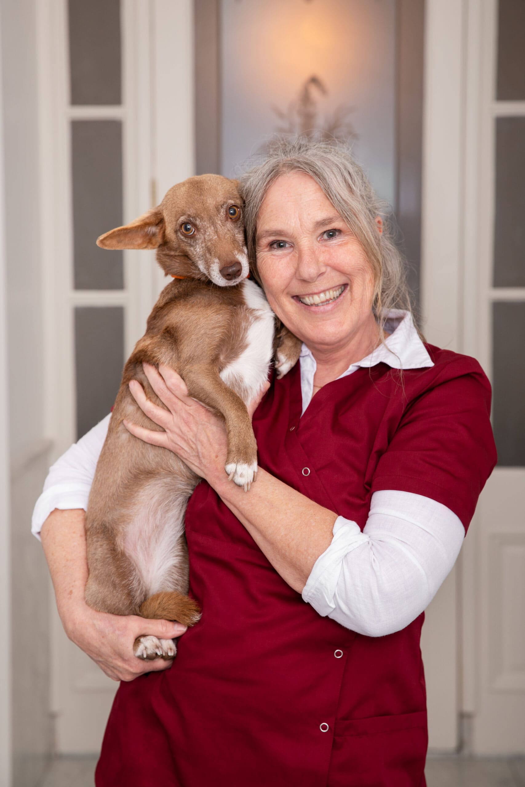 Medewerker in bordeauxrode dienstkleding met een kleine bruin met witte hond op de arm.